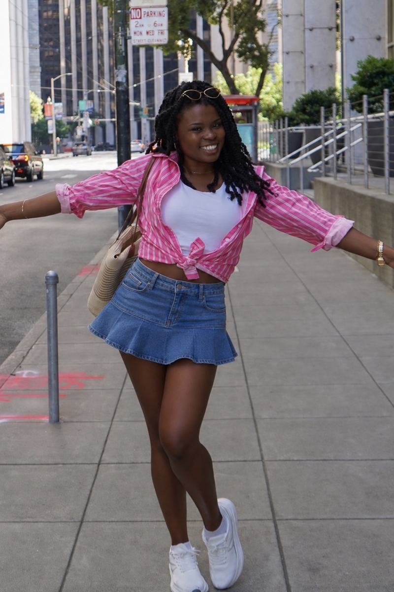 Faye Constant walking a city street in a pink shirt and denim skirt, arms open